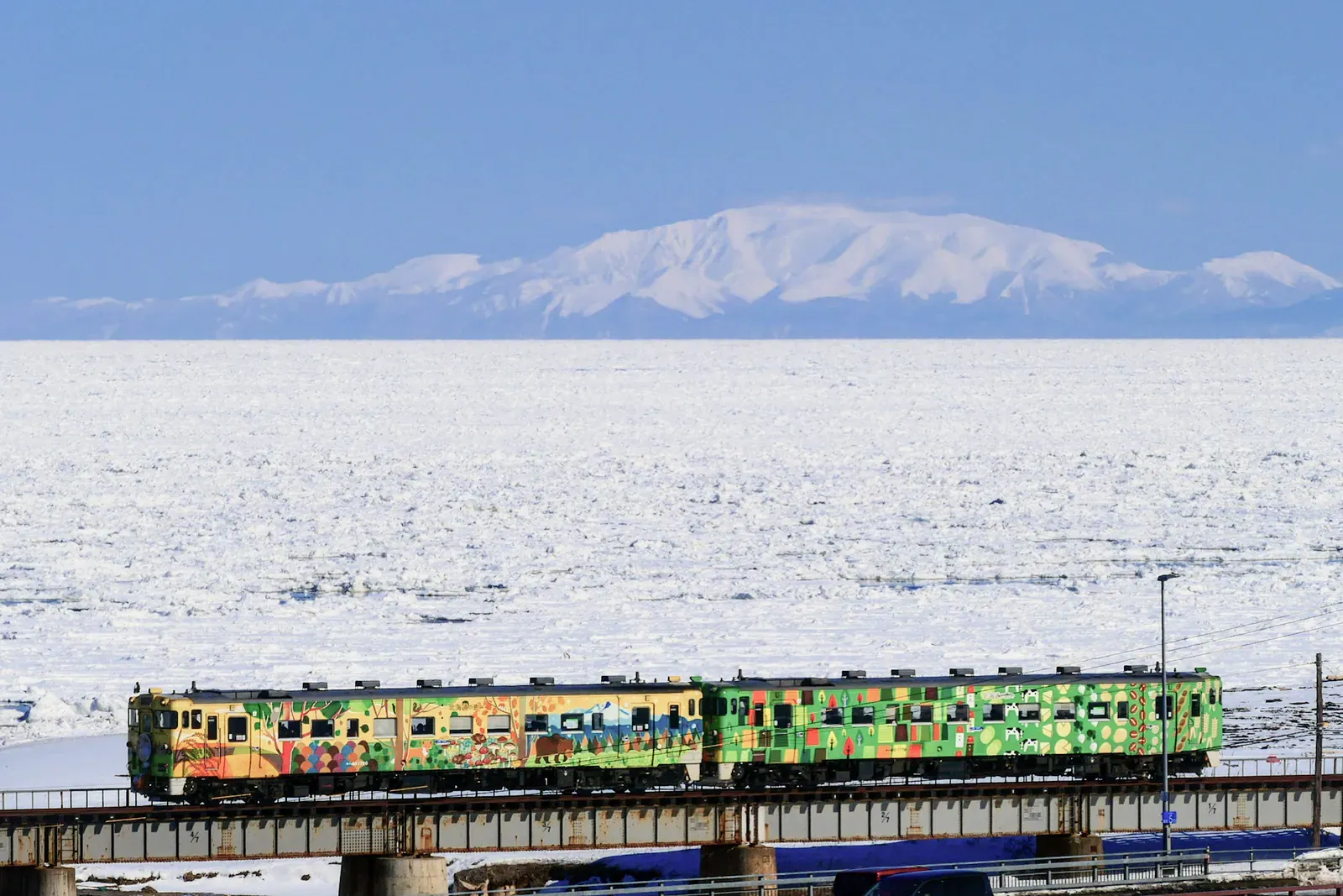 Colourful two-car train on a bridge before a frozen sea and white mountains